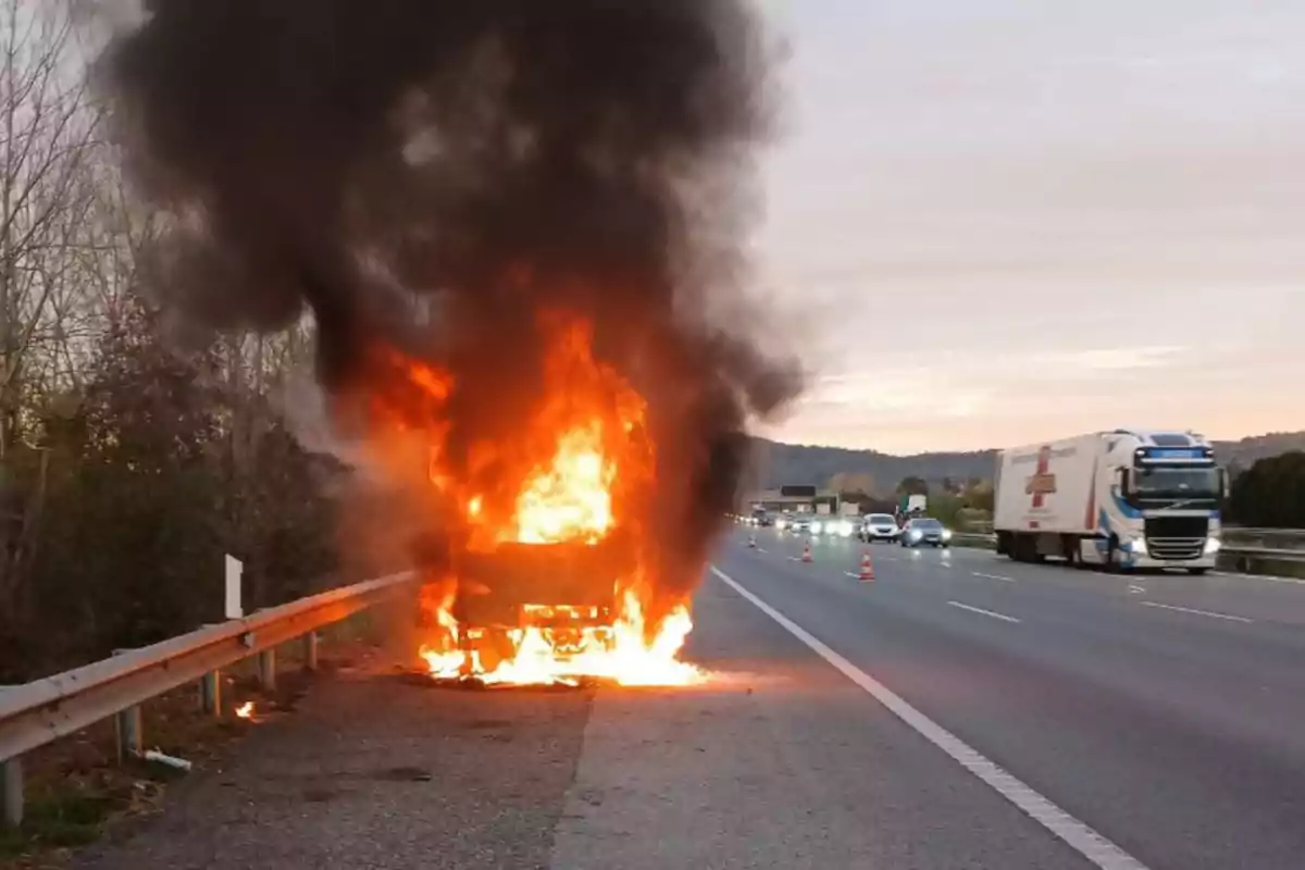 Un vehículo en llamas en el arcén de una autopista con humo negro elevándose al cielo mientras los coches pasan por el carril adyacente.