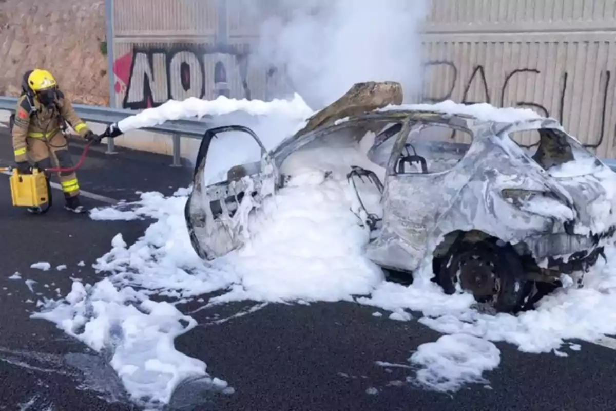 Un bombero apaga un coche incendiado con espuma en una carretera.