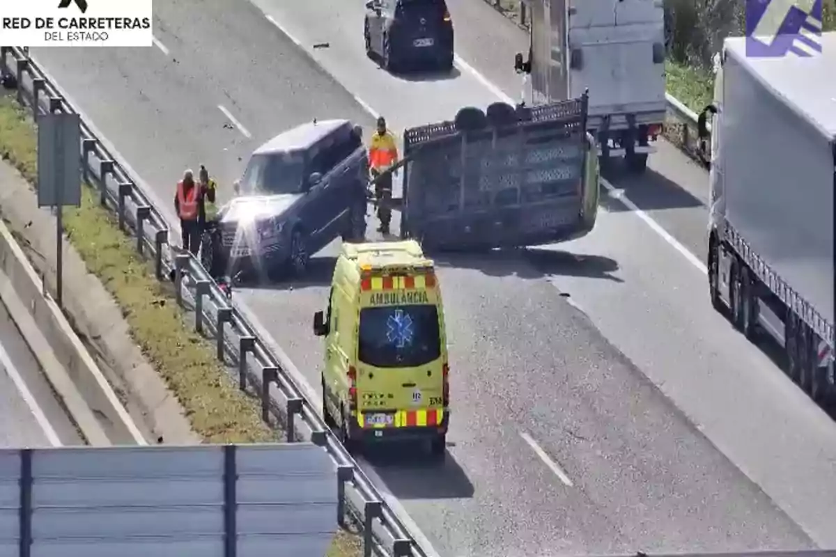 Una ambulancia y varios vehículos están en una carretera donde un remolque ha volcado, con personas alrededor evaluando la situación.