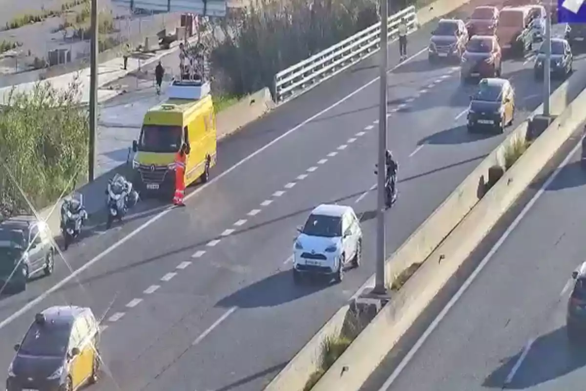 Una carretera con tráfico moderado donde se observa una furgoneta amarilla detenida en el arcén junto a dos motocicletas de policía y varios coches circulando.