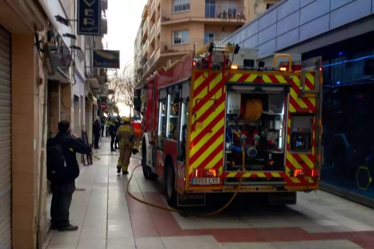 Un camió de bombers estacionat en un carrer estret mentre els bombers treballen al lloc.