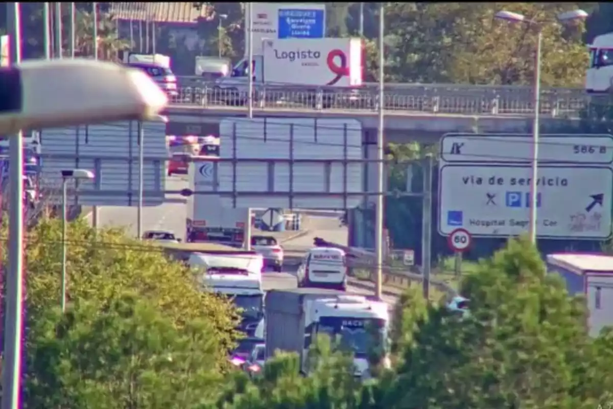 Una carretera amb trànsit dens i diversos vehicles circulant en totes dues direccions, amb un pont elevat i senyals de trànsit visibles.