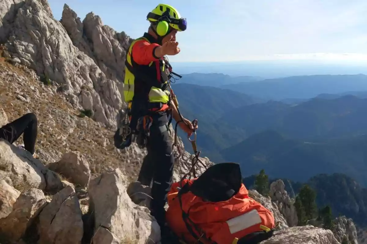 Un rescatista equipat amb casc i arnès es troba en una muntanya rocosa al costat d´una llitera taronja, amb un paisatge muntanyós al fons.