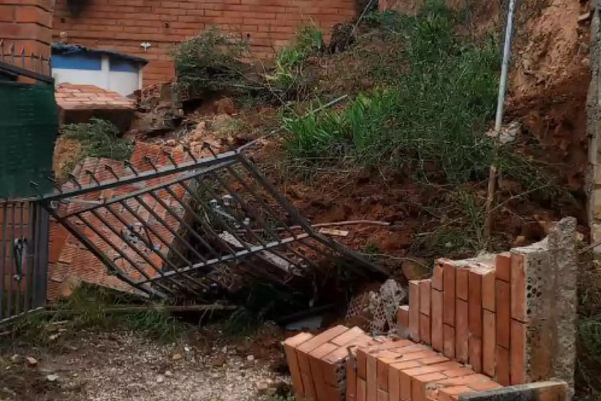 Una reixa de metall caiguda sobre un munt de maons i terra en una àrea amb vegetació i un mur de maó al fons.