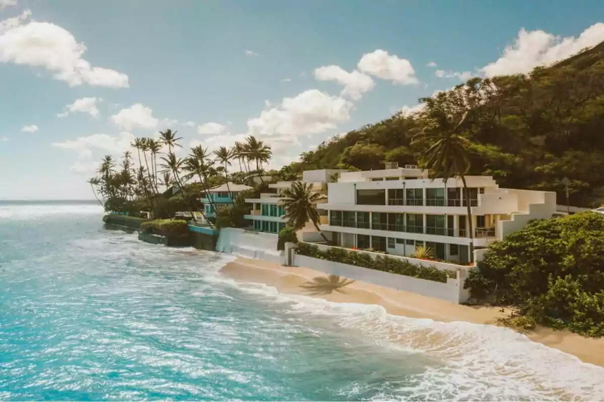 Vista aérea de una casa moderna frente al mar con palmeras y una playa de arena dorada bajo un cielo azul con nubes dispersas.