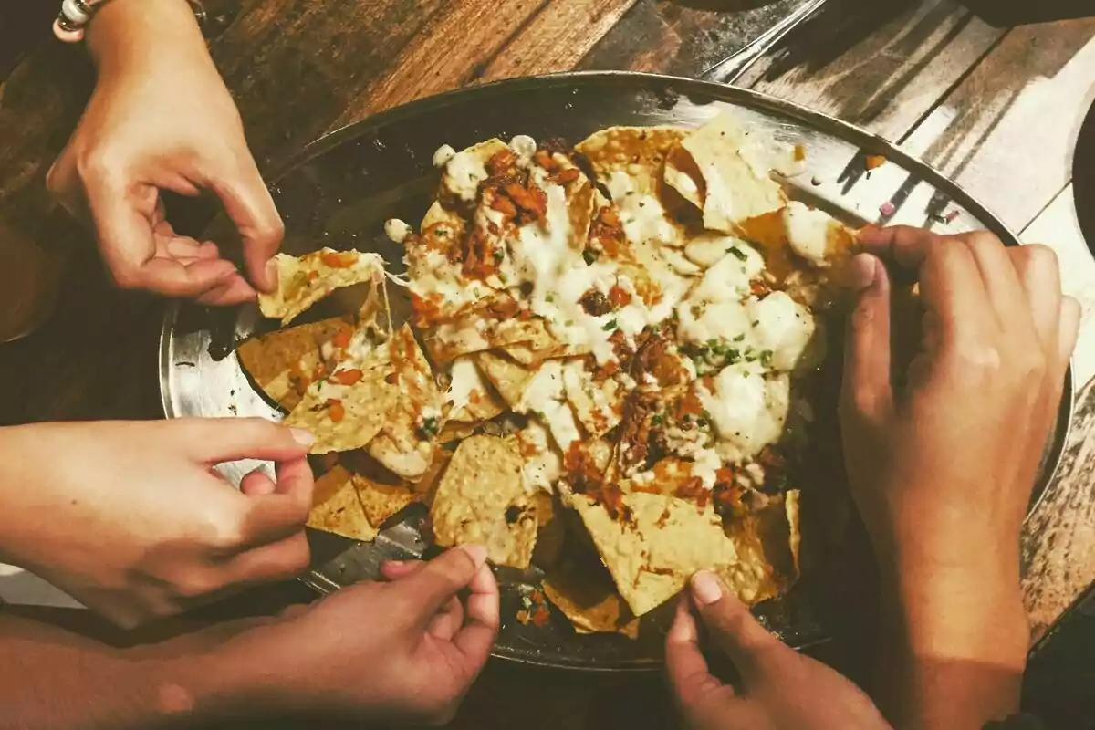 Varias manos tomando nachos con queso derretido de un plato grande sobre una mesa de madera.