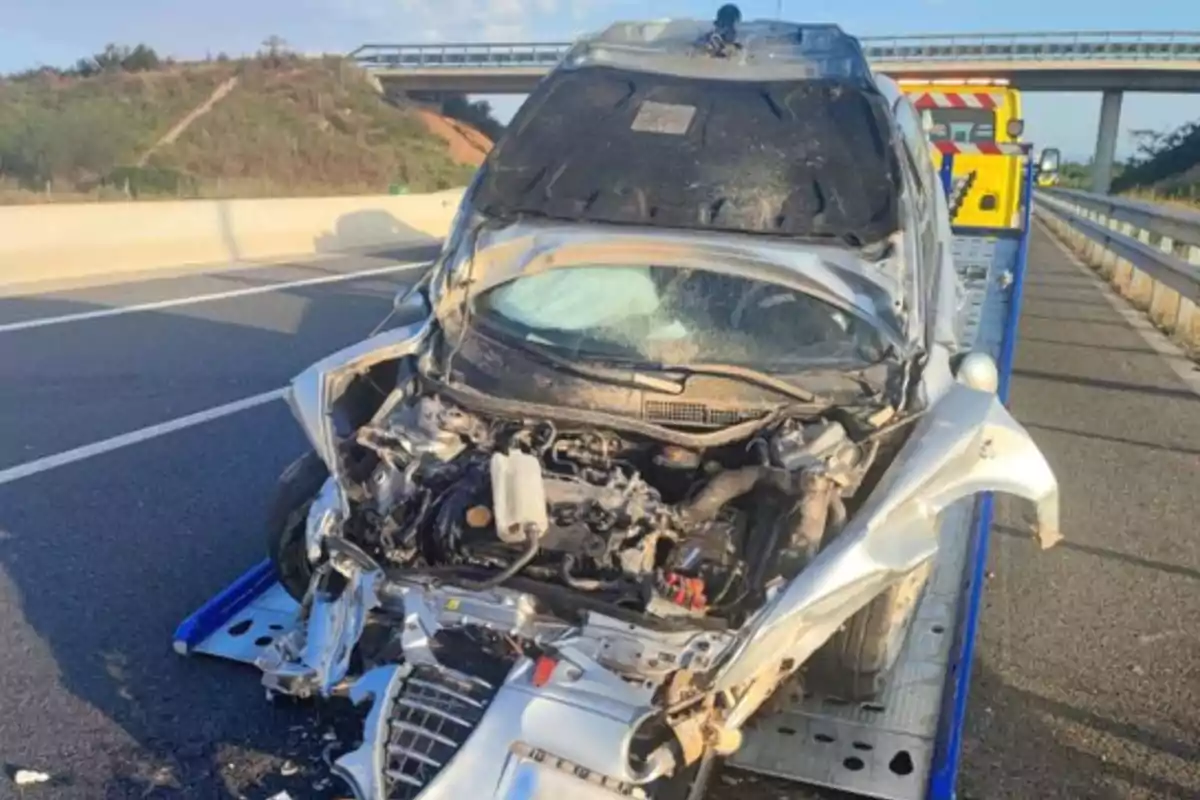 Un cotxe platejat amb la part davantera greument malmesa està sent remolcat en una grua en una carretera.