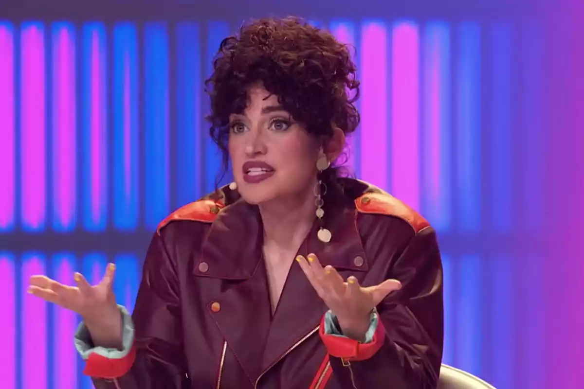 A woman with dark curly hair, wearing a brown leather jacket with red details and long earrings, gestures as she speaks, against a background of blue and pink lights.