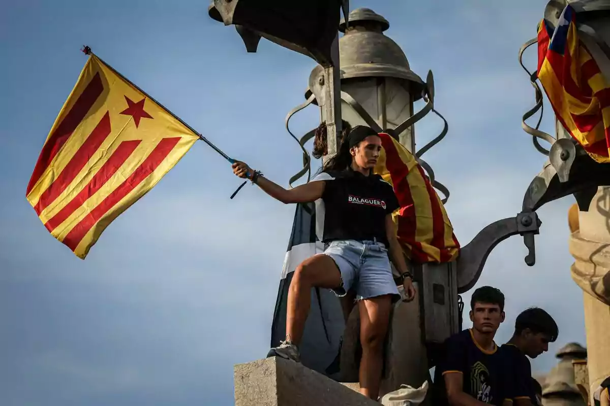 Una dona sosté una bandera amb ratlles vermelles i grogues i una estrella vermella mentre està dreta sobre una estructura amb dos joves.