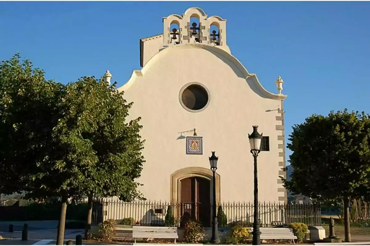 Fachada de una iglesia blanca con campanario y árboles a los lados bajo un cielo azul.