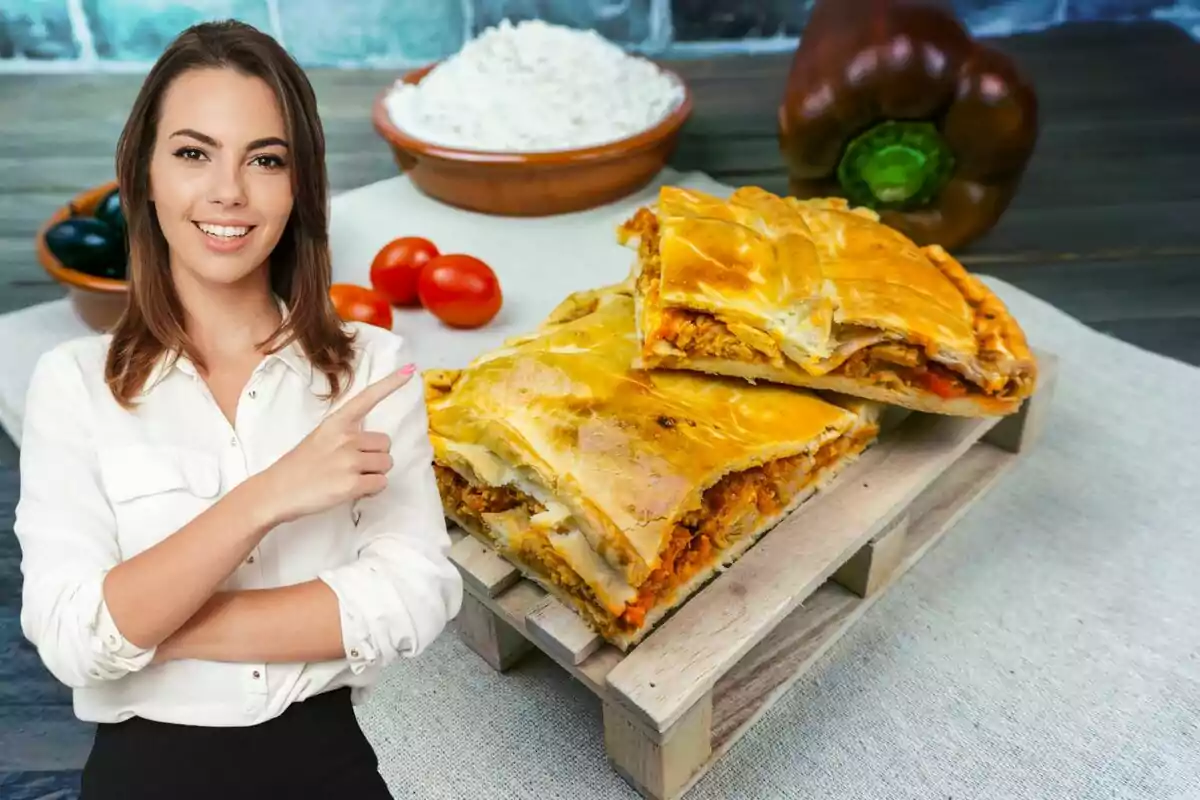 Una dona somrient amb camisa blanca assenyala cap a una empanada gallega sobre una taula, amb ingredients com tomàquets cherry, pebrots i farina al fons.