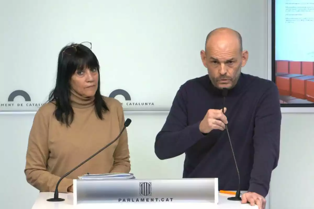 Dues persones en una conferència de premsa al Parlament de Catalunya, una dona amb jersei marró i un home amb jersei fosc, tots dos davant de micròfons.