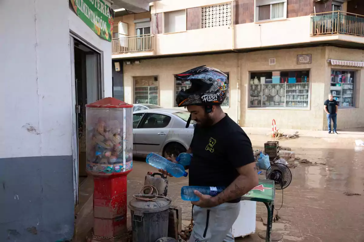 Un home amb casc sosté ampolles d'aigua davant d'una botiga en un carrer amb fang després de la DANA