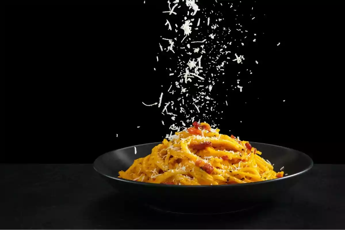 Un plato de pasta con queso rallado cayendo sobre él, en un fondo negro.