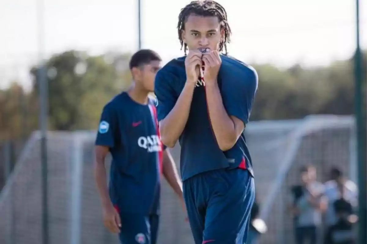 Dos jugadors de futbol amb uniforme blau en un camp d'entrenament, un besa l'escut de la samarreta.