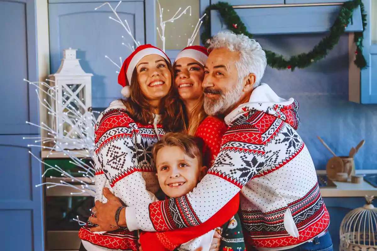 Una família somrient amb suèters nadalencs i gorres de Santa Claus s'abraça en una cuina decorada pel Nadal.