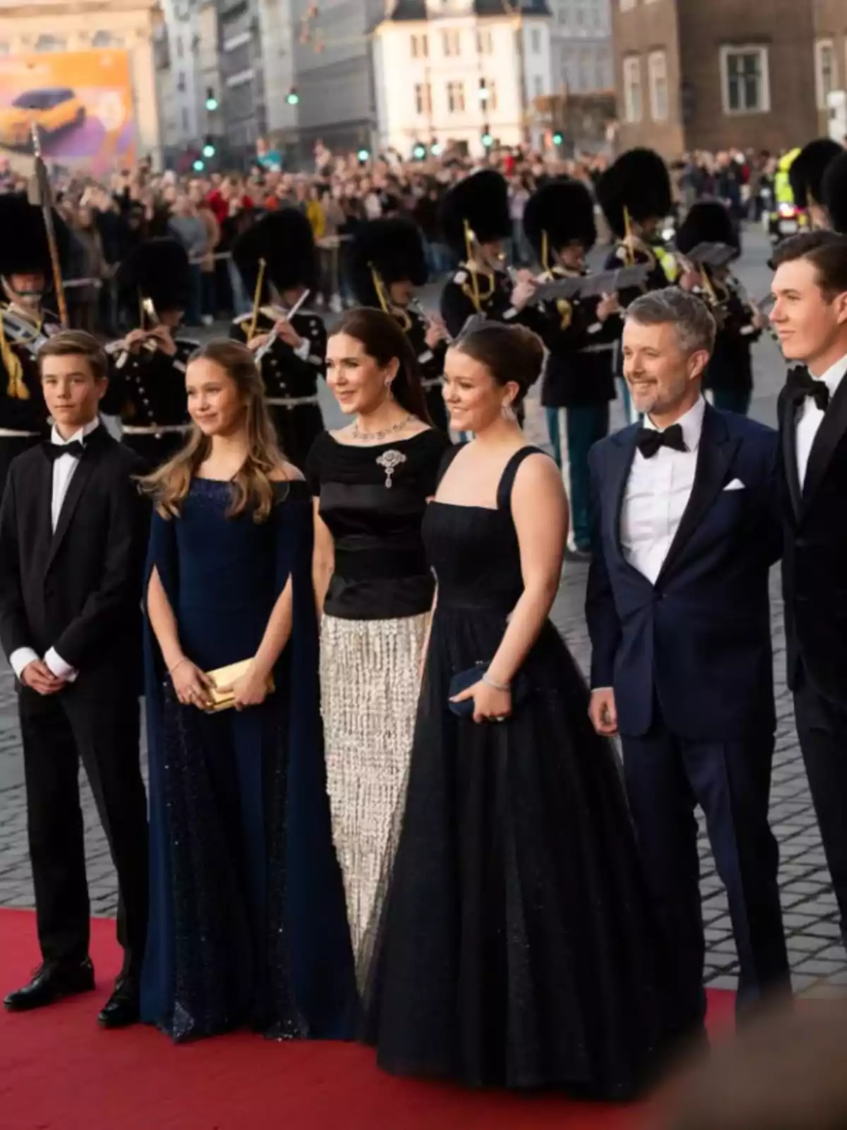 A group of elegantly dressed people poses on a red carpet while a band of musicians in uniforms and tall hats plays in the background, surrounded by a crowd of spectators in an urban setting. A group of elegantly dressed people poses on a red carpet while a band of musicians in uniforms and tall hats plays in the background, surrounded by a crowd of spectators in an urban setting.