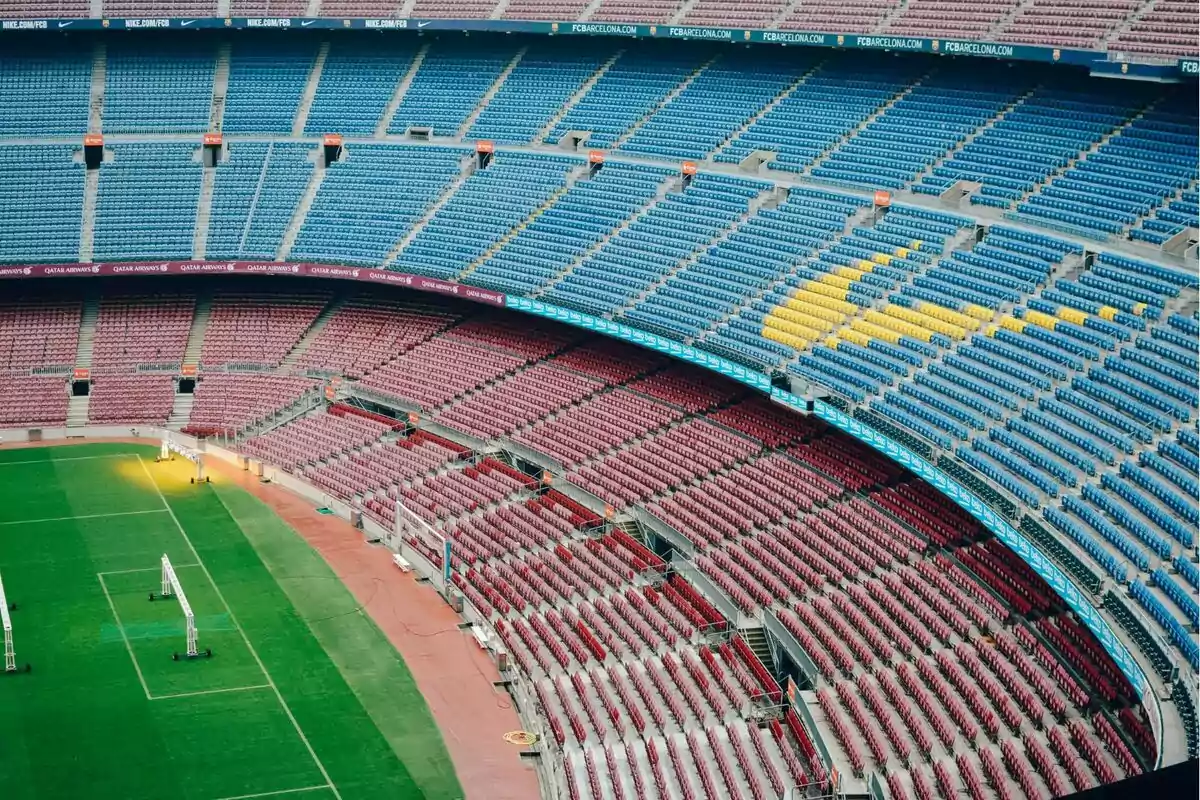 Vista interior d´un estadi de futbol buit amb grades de colors vermell, blau i groc.