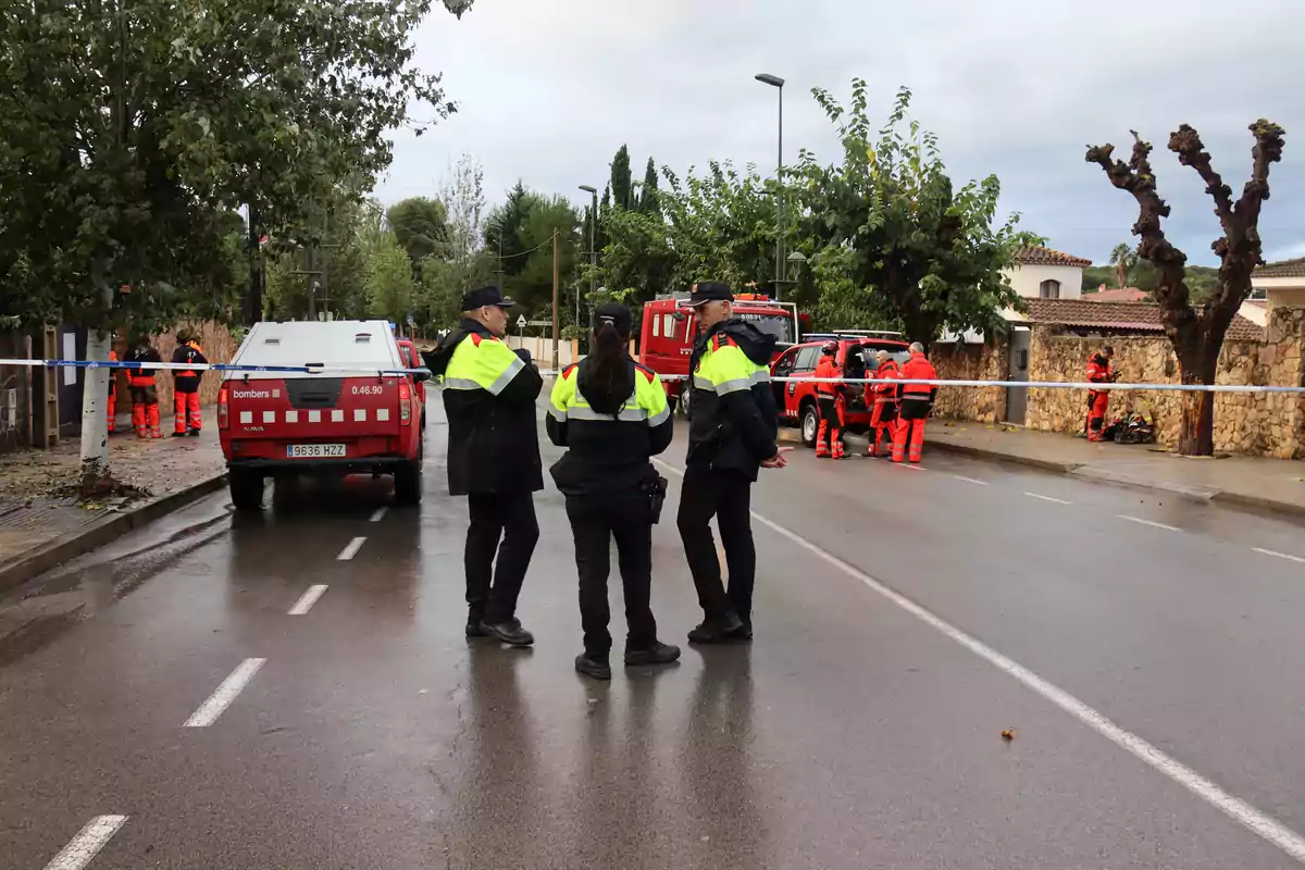 Un grupo de bomberos y personal de emergencia en una calle mojada con vehículos de servicio y cinta de seguridad.