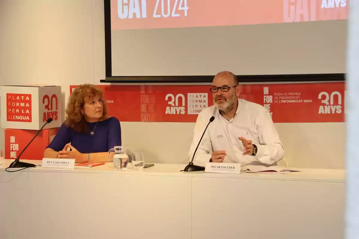 Dues persones assegudes en una taula durant una conferència de premsa de la Plataforma per la Llengua, amb un cartell que diu “30 anys” i “CAT 2024” al fons. Dues persones assegudes en una taula durant una conferència de premsa de la Plataforma per la Llengua, amb un cartell que diu “30 anys” i “CAT 2024” al fons.