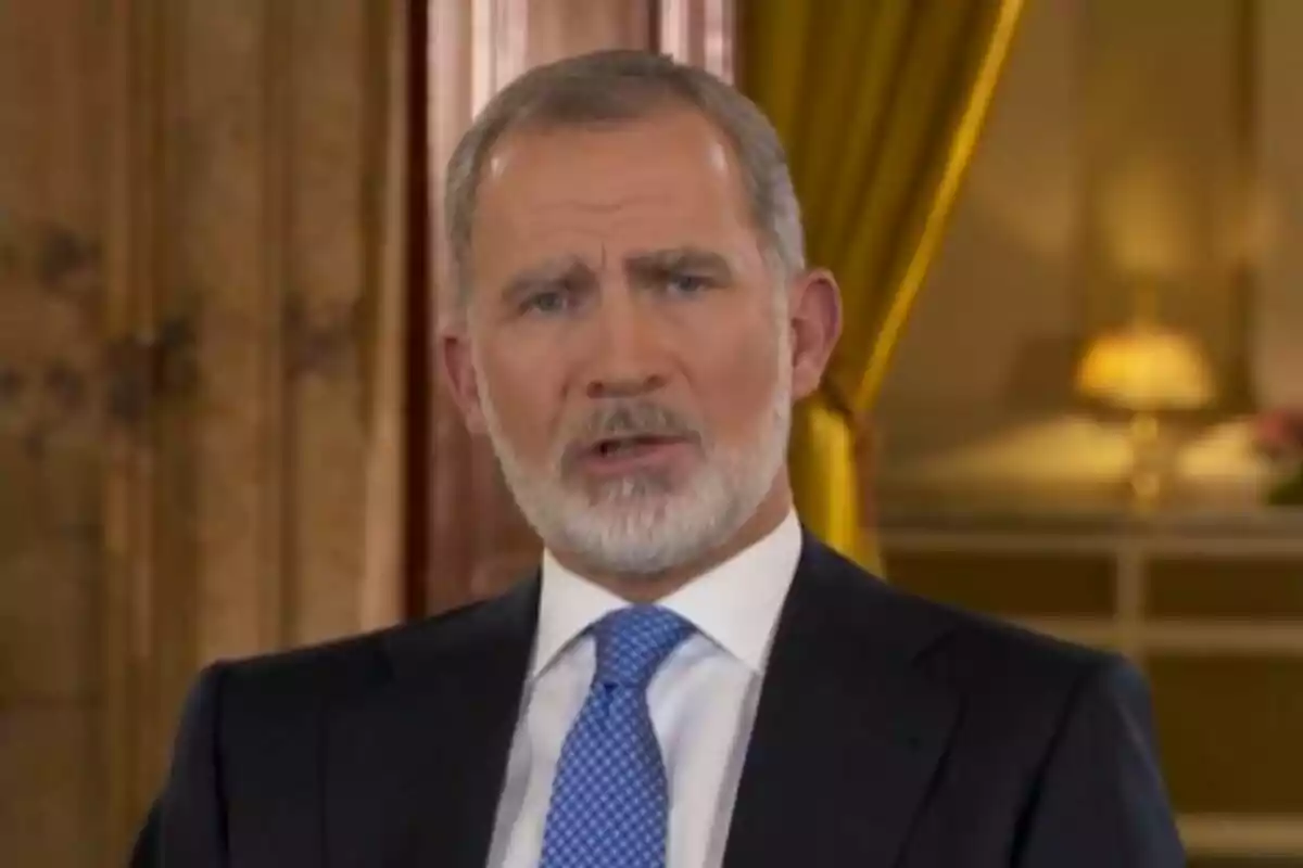 Un hombre con barba y traje azul aparece en un entorno elegante con cortinas amarillas y una lámpara de fondo.