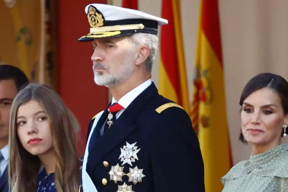 Un hombre con uniforme militar y gorra blanca está acompañado por dos mujeres, una joven y una adulta, con banderas españolas de fondo.