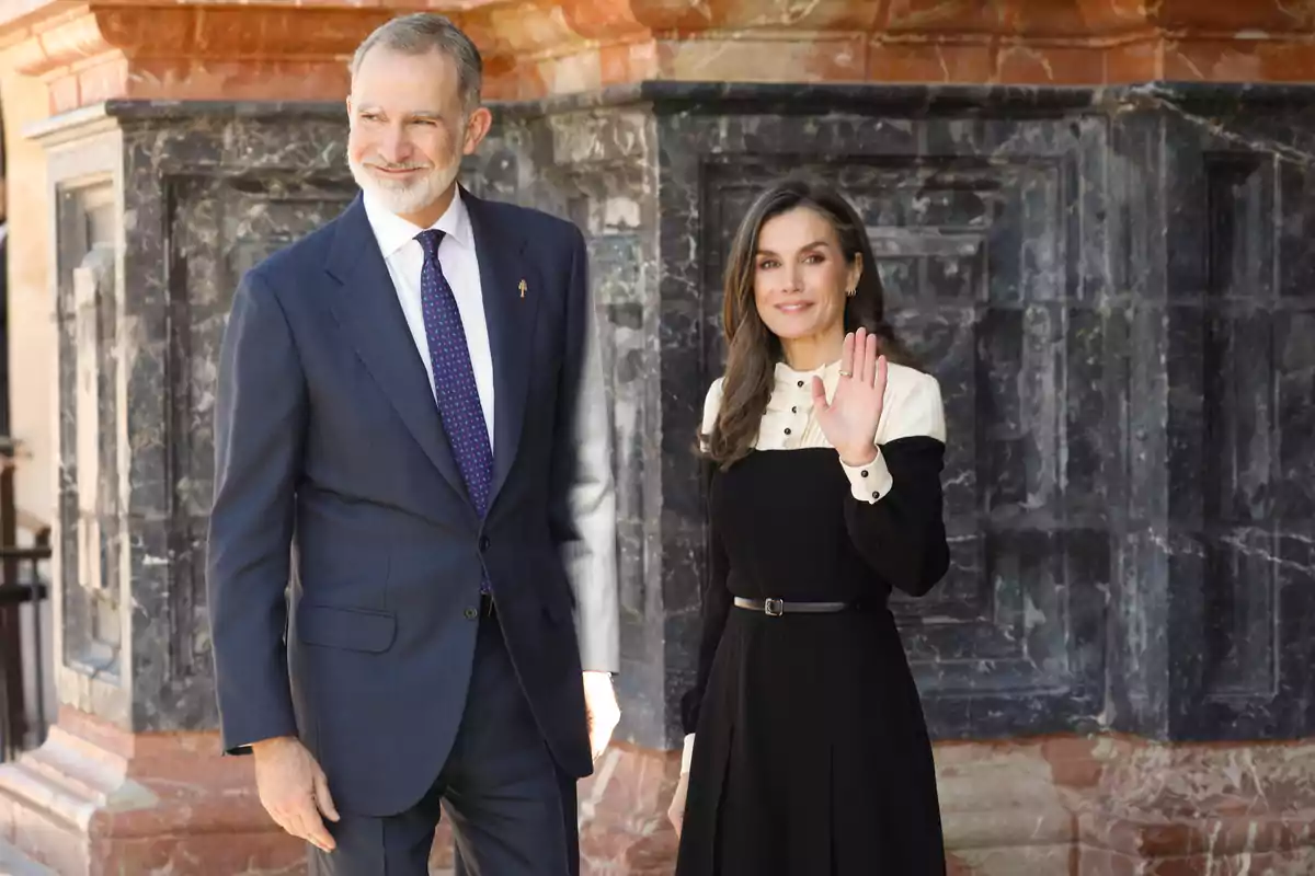Felipe VI y Letizia vestida formalmente, el hombre lleva un traje oscuro y la mujer un vestido negro con detalles claros, ambos están de pie frente a una estructura de piedra. Felipe VI y Letizia vestida formalmente, el hombre lleva un traje oscuro y la mujer un vestido negro con detalles claros, ambos están de pie frente a una estructura de piedra.