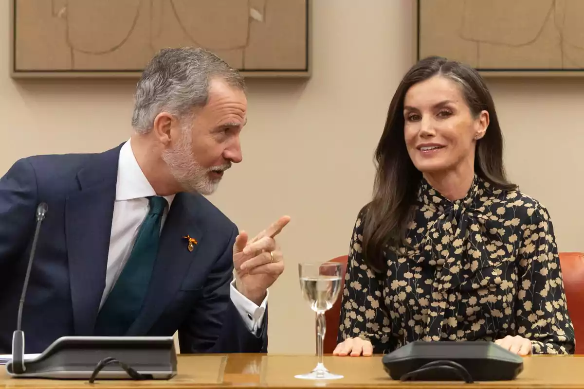 Felip VI i la reina Letícia asseguts a una taula conversant, una d'elles gesticula mentre l'altra somriu, amb un micròfon i una copa d'aigua davant d'ells. Felip VI i la reina Letícia asseguts a una taula conversant, una d'elles gesticula mentre l'altra somriu, amb un micròfon i una copa d'aigua davant d'ells.