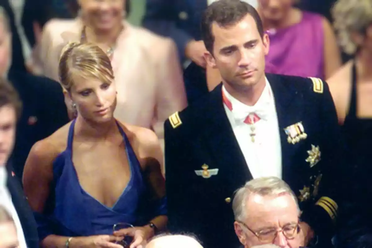 Una pareja elegantemente vestida en un evento formal rodeada de otras personas.