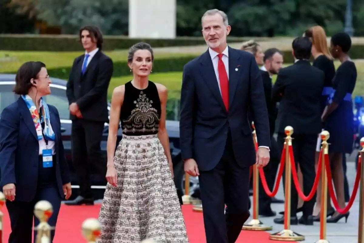 Una parella elegantment vestida camina per una catifa vermella, envoltada d'altres persones i cordes de vellut vermell.