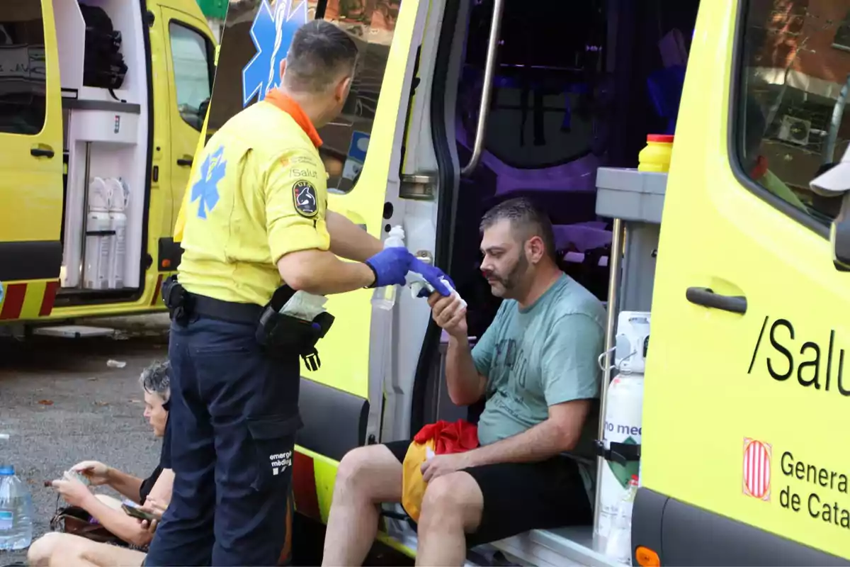 Un paramèdic atén un home assegut a la porta d'una ambulància mentre una altra persona està asseguda a terra a prop seu.