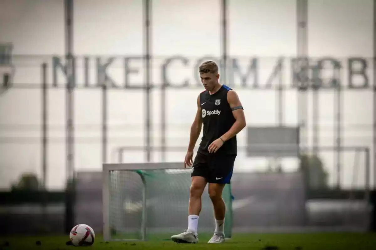 Un jugador de futbol amb uniforme d'entrenament negre i una pilota al camp de joc, amb una porteria i un cartell de Nike al fons.