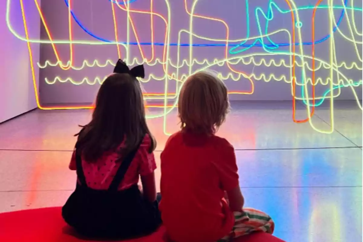 Dos nens asseguts a terra observen una instal·lació de llums de neó de colors brillants en una galeria d'art. Dos nens asseguts a terra observen una instal·lació de llums de neó de colors brillants en una galeria d'art.
