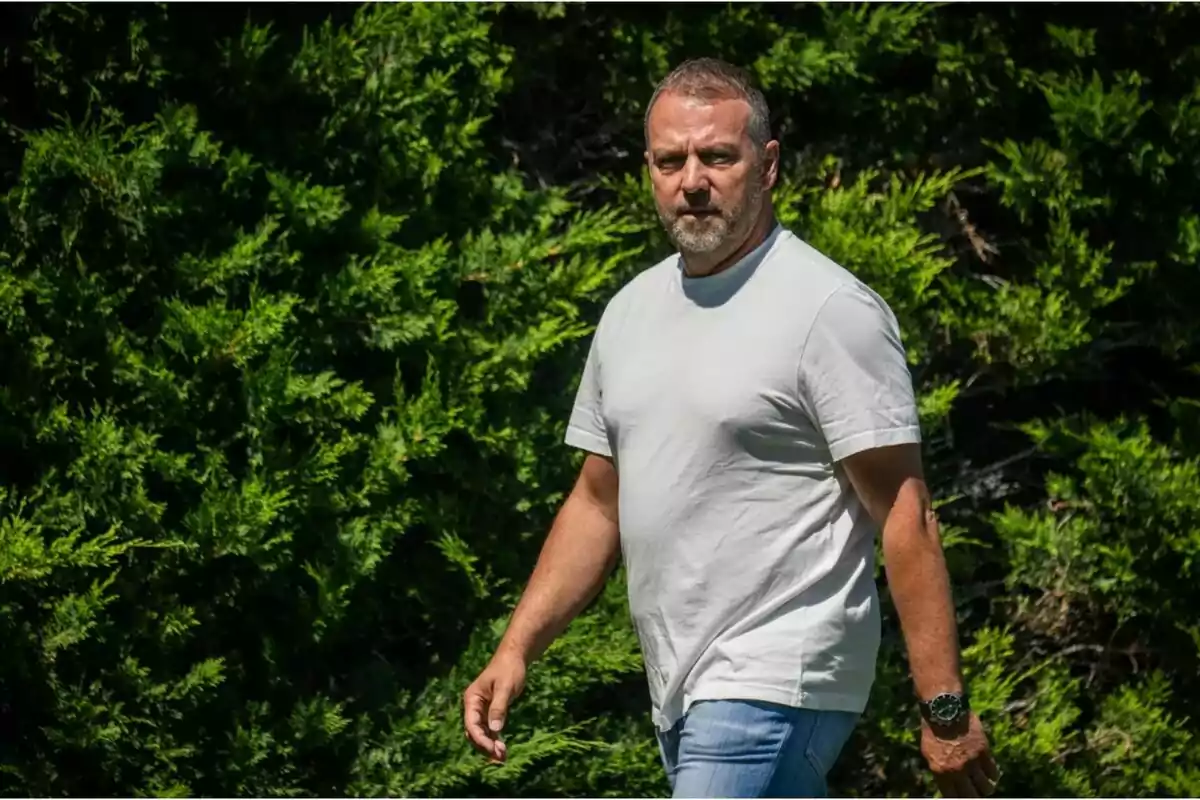 Hombre con camiseta blanca y jeans caminando al aire libre frente a un fondo de arbustos verdes.