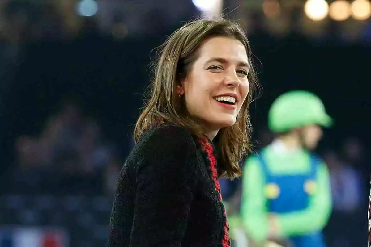 Charlotte Casiraghi smiling with brown hair and a dark sweater in a setting with bright lights and a person in costume in the background. Charlotte Casiraghi smiling with brown hair and a dark sweater in a setting with bright lights and a person in costume in the background.