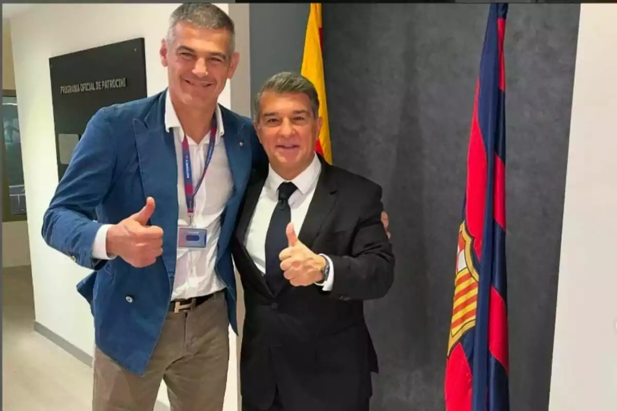 Dos hombres sonrientes posan juntos con pulgares arriba frente a una bandera y un cartel que dice "Programa Oficial de Patrocinio".