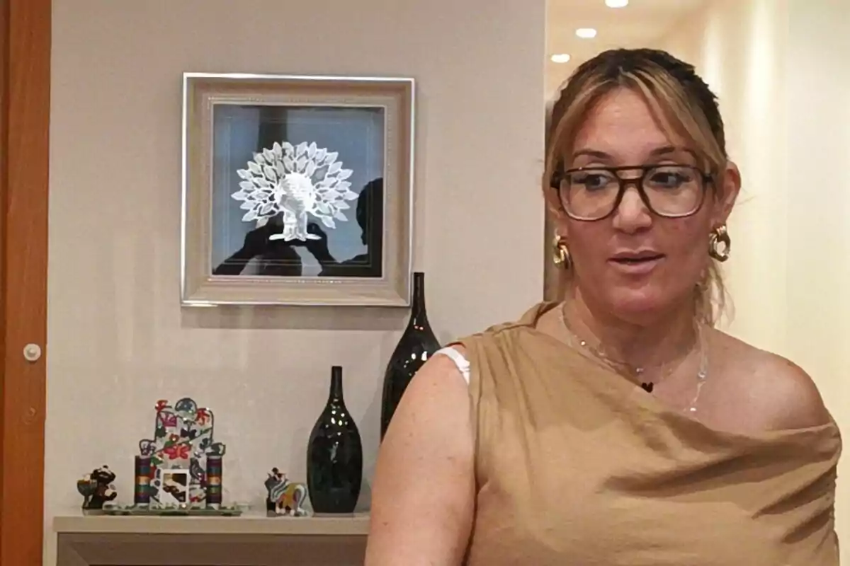 Una dona amb ulleres i arracades daurades està dreta davant d'una paret decorada amb un quadre d'un arbre i diversos objectes acolorits sobre una lleixa.