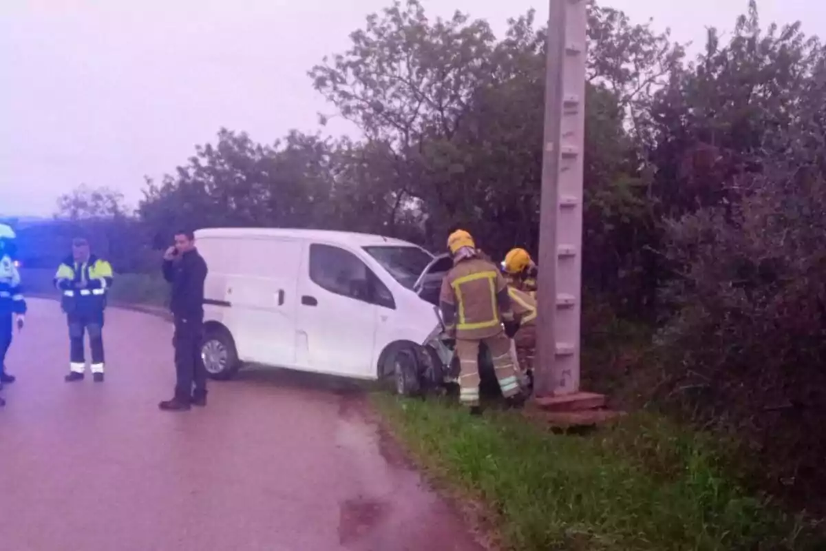 Una furgoneta blanca ha xocat contra un pal en una carretera rural mentre bombers i personal d'emergència avaluen la situació.