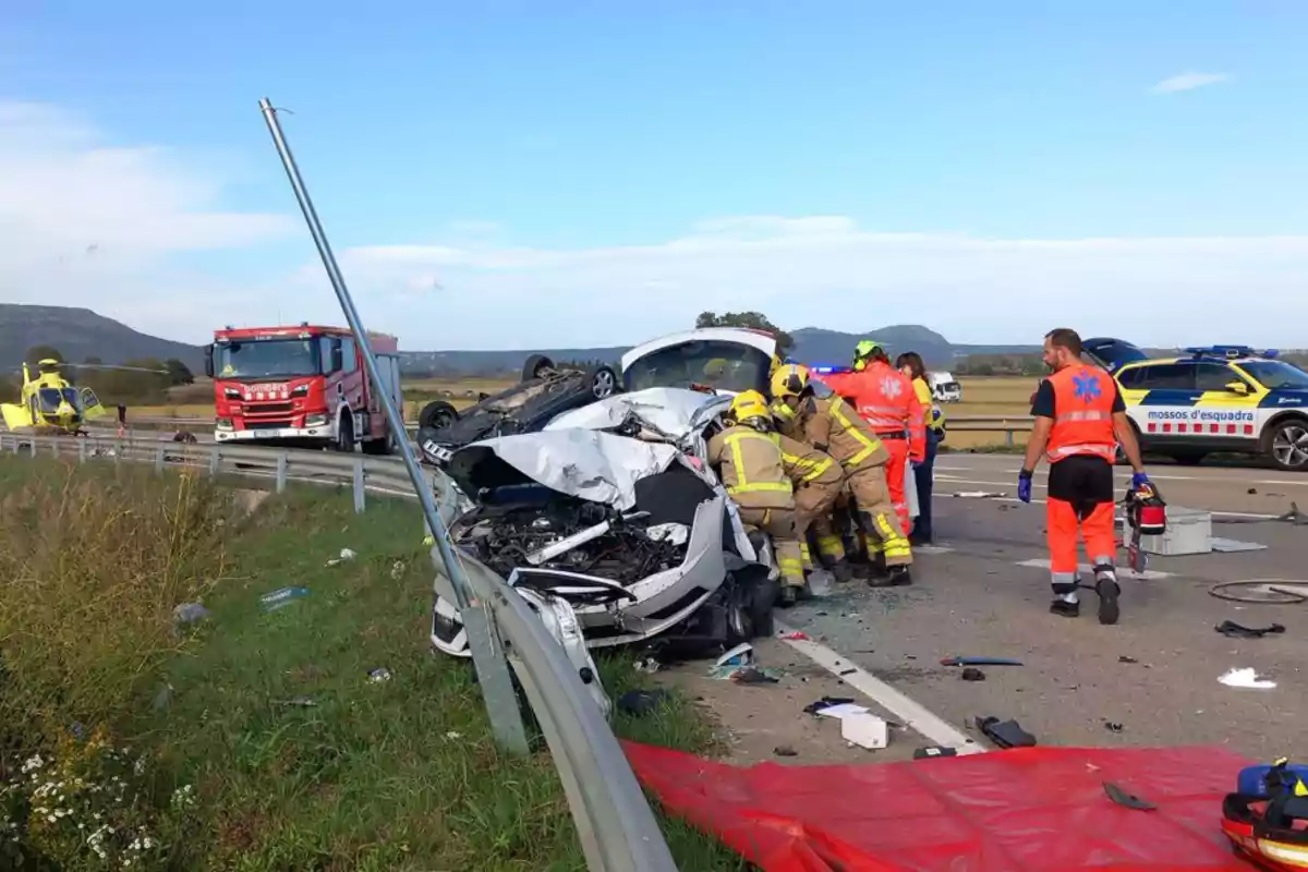 Accident de trànsit en una carretera amb un cotxe bolcat i un altre greument danyat; bombers i personal d'emergència treballant al lloc, amb un camió de bombers, un helicòpter i un cotxe de policia presents.