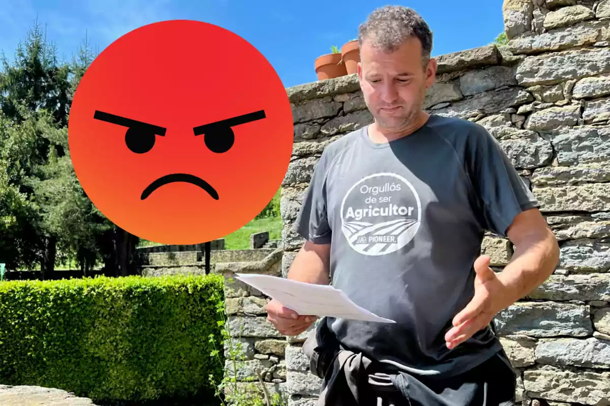 Home amb samarreta de pagès sostenint papers al costat d'un emoji d'enfadament en un entorn a l'aire lliure amb murs de pedra i vegetació.
