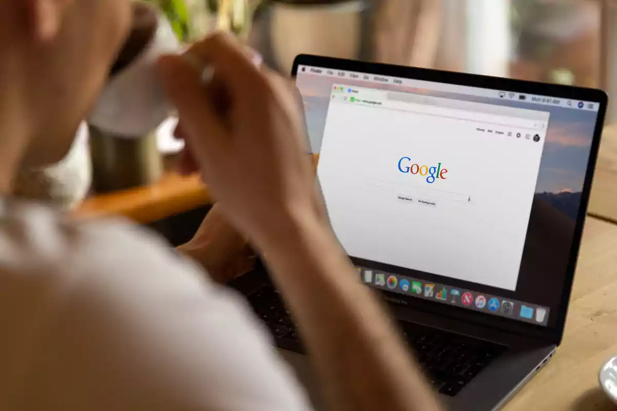 Persona usando una laptop con la página de inicio de Google abierta mientras sostiene una taza de café.