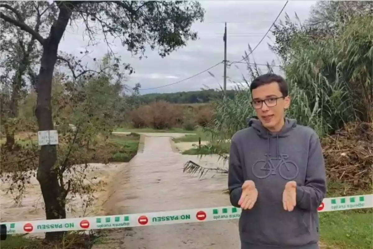 Un joven con sudadera gris está de pie frente a un camino inundado que está bloqueado con una cinta de advertencia que dice "NO PASAR" rodeado de árboles y vegetación.