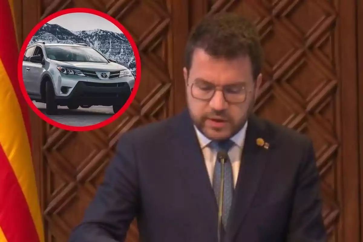 Un home amb ulleres i vestit fosc parla en un podi, mentre a la cantonada superior esquerra es mostra la imatge d?un cotxe Toyota emmarcat en un cercle vermell.