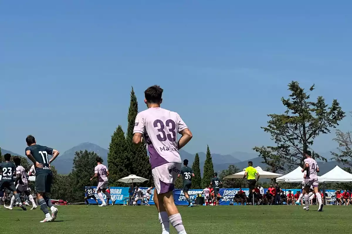 Jugadors de futbol en un camp durant un partit amb muntanyes i arbres al fons.