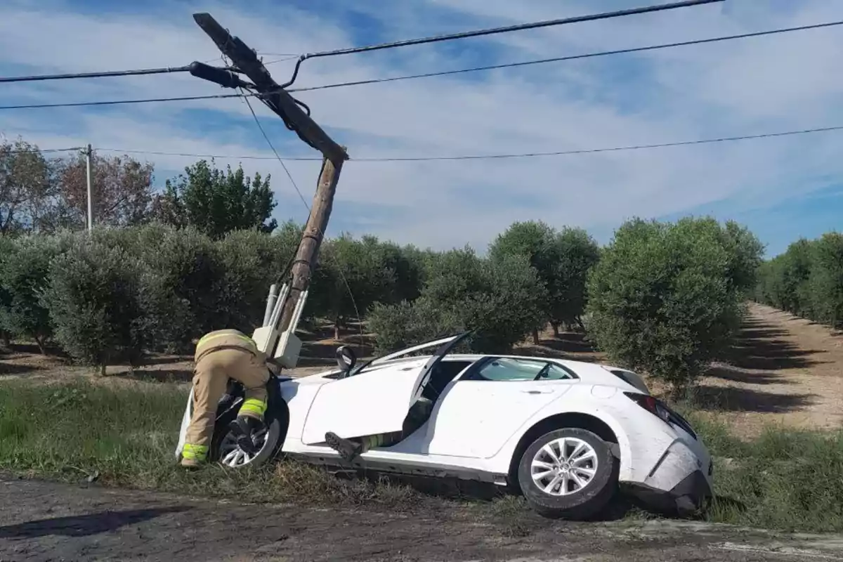 Un cotxe blanc xoca contra un pal d'electricitat a una carretera rural mentre un bomber inspecciona el mal.