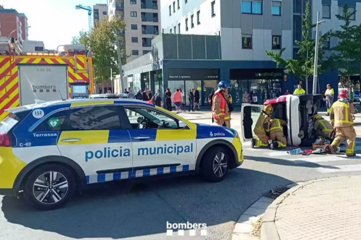 Un cotxe de la policia municipal i un camió de bombers al?escena d?un accident de trànsit amb un vehicle bolcat i bombers assistint.