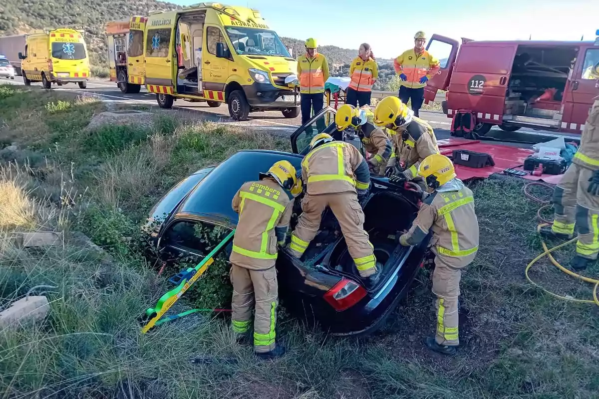 Bombers i personal d'emergència treballant en el rescat d'un cotxe accidentat en una rasa amb ambulàncies i vehicles d'emergència al fons.