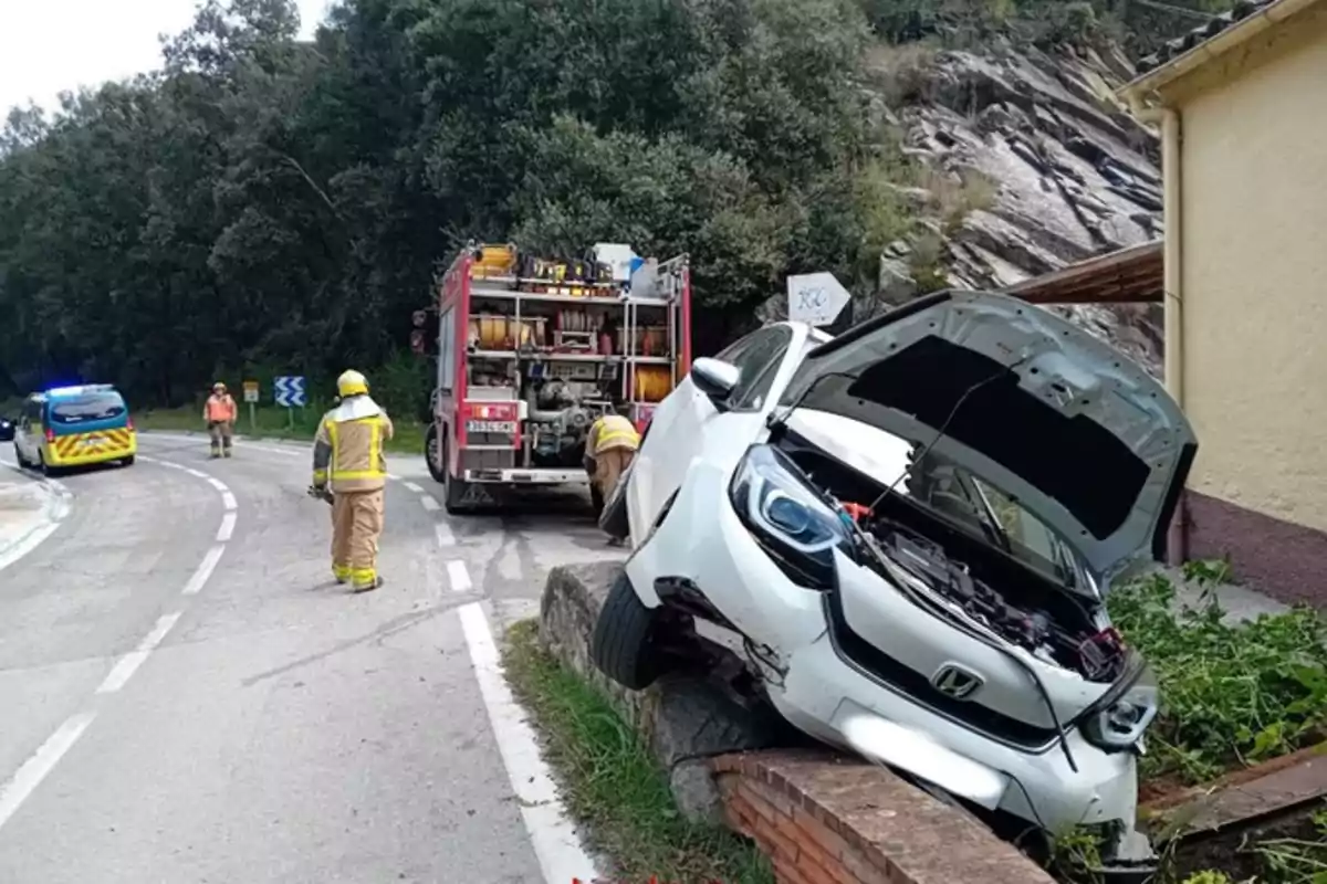 Un cotxe blanc accidentat sobre un mur al costat d?una carretera amb un camió de bombers i personal d?emergència al?escena.