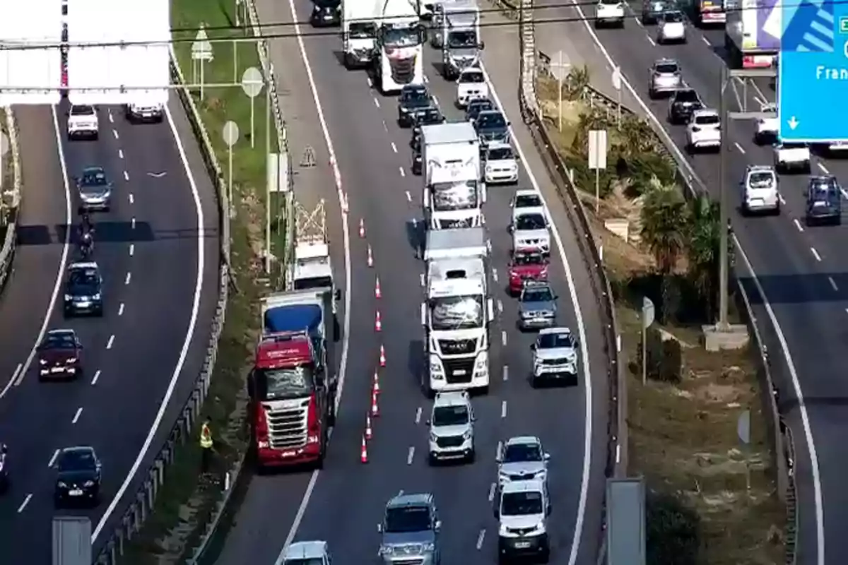 Una carretera con tráfico denso donde se observan varios vehículos, incluyendo camiones y coches, circulando en ambas direcciones, con conos naranjas delimitando un carril.