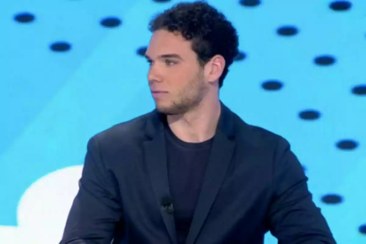 Un hombre con cabello rizado y barba ligera, vestido con un saco oscuro, está sentado frente a un fondo azul con puntos negros.